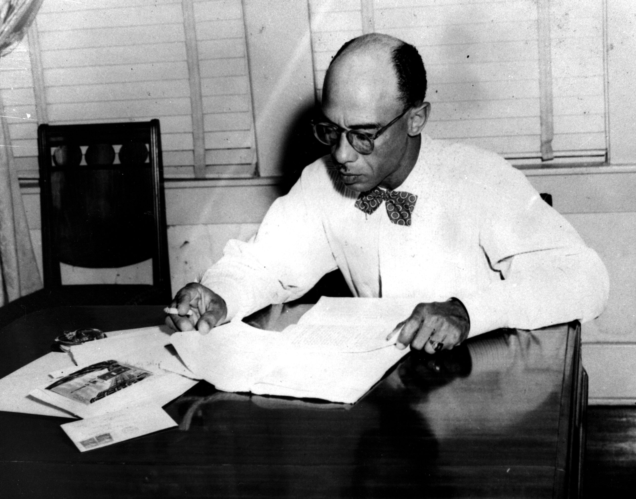 A balding African American man in a bow tie studies documents at a wooden desk