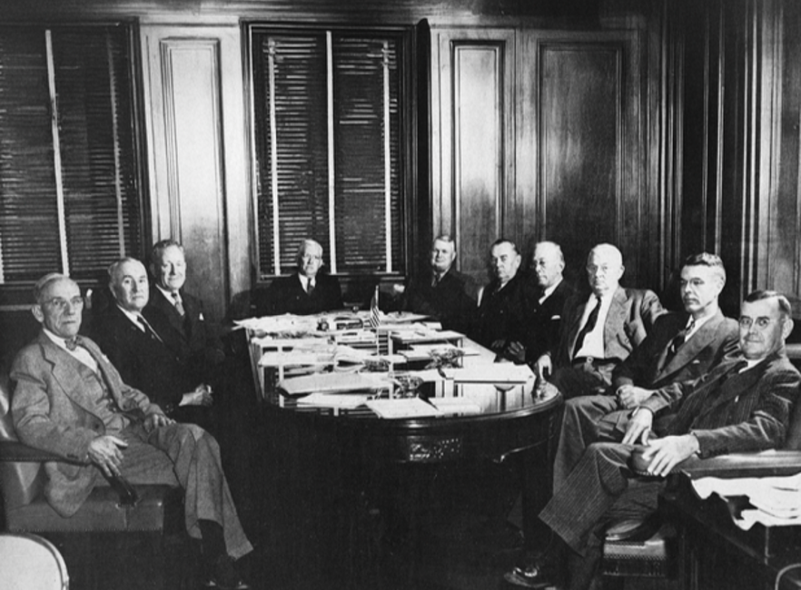 Ten elderly white men in executive business attire from the 1950s South sit for a portrait around a conference table in a wood paneled board room.