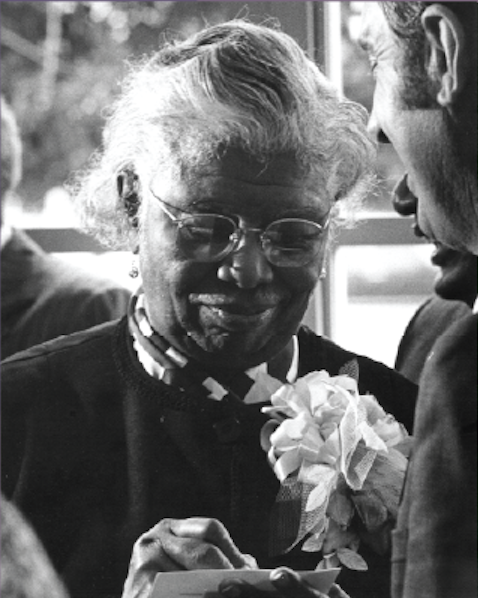 An elderly woman in church clothes wearing a corsage writes a note on a card