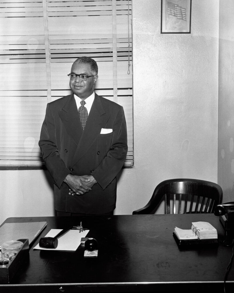 A man in business attire stands behind a desk with his hands folded in front of him