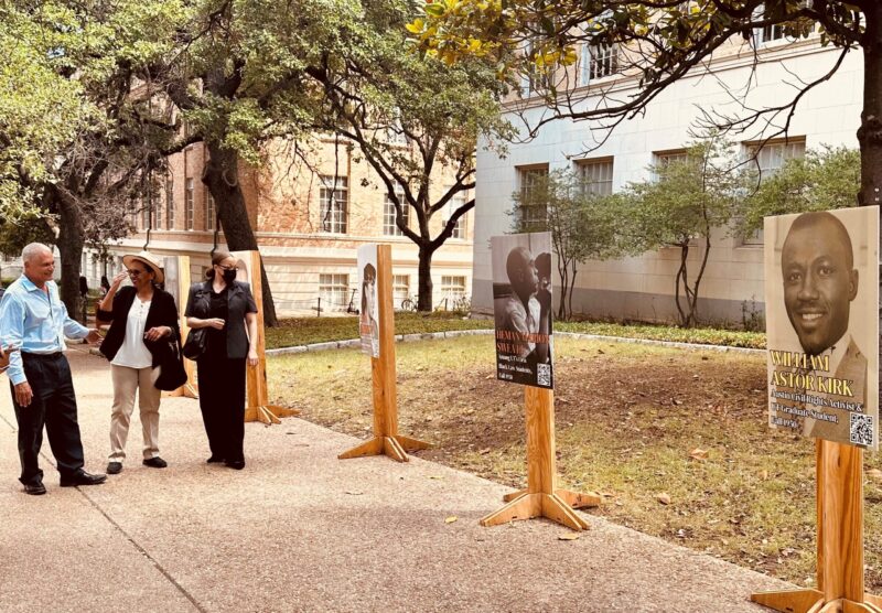 Three college professors stand on a campus sidewalk beside a display of three vinyl posters mounted on stands which are part of an outdoor history exhibit.
