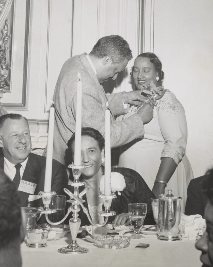 An elderly man pins a corsage on the shoulder of an elderly woman in a banquet hall