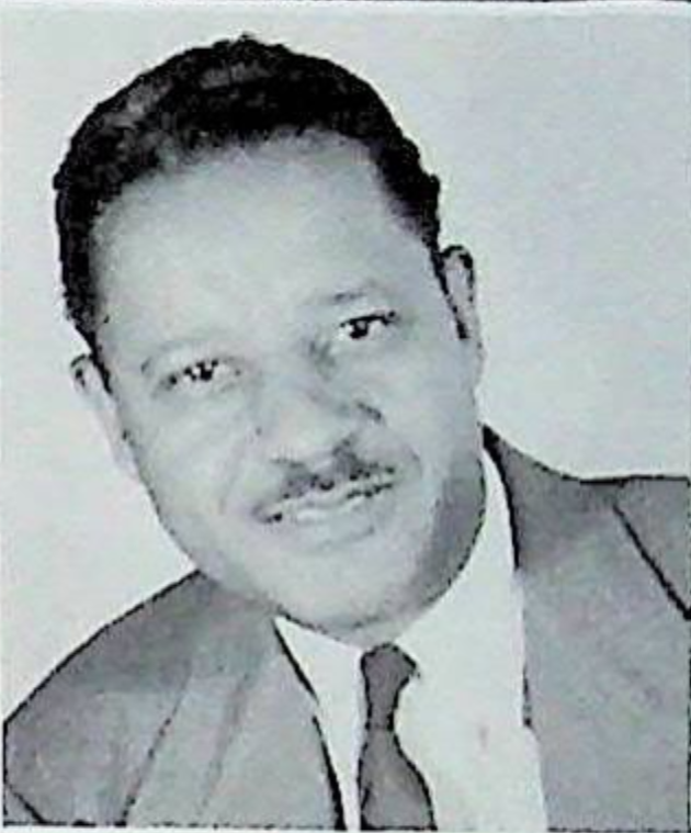 A mustached man in a suit and tie looks at the camera