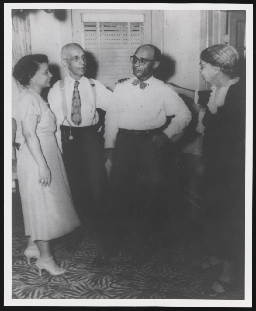 Four adults, including Heman Sweatt, in formal attire stand in a circle conversing