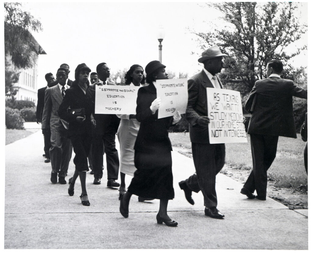 Nine African American adults in business attire and holding placards march on a sidewalk
