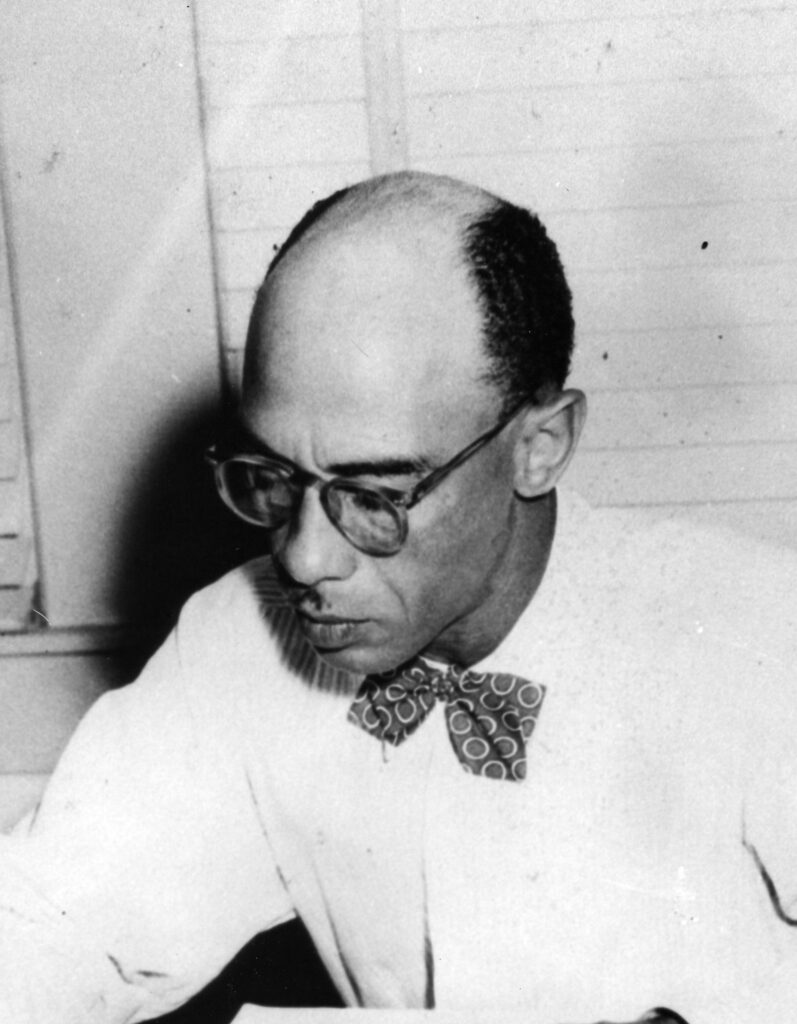 A balding man in glasses, a dress shirt and a bow tie studies something on a desk out of frame