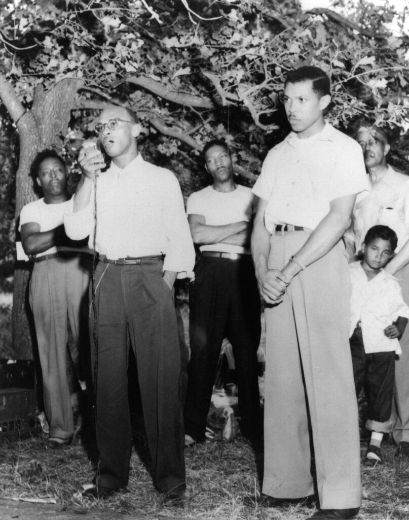 Two men stand in the foreground in slacks and dress shirts. One man is holding a microphone. A tree and a small group of people stand a few feet behind them.