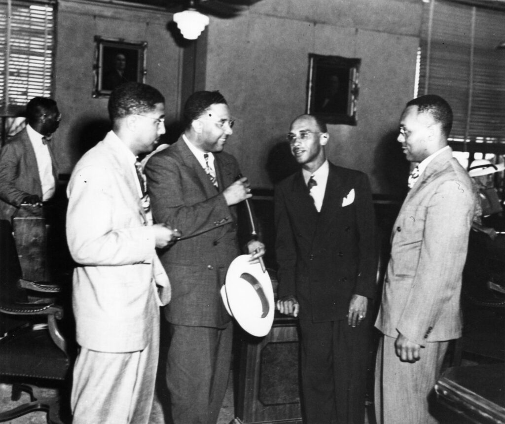 Four African American men in business attire stand in a courtroom conversing among themselves