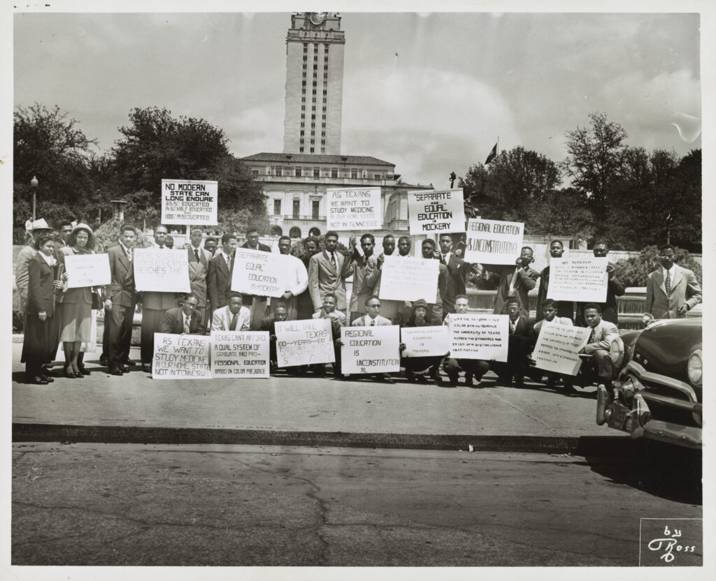 A small crowd of protesters holding placard sit on the steps in front of the Tower building on UT Campus