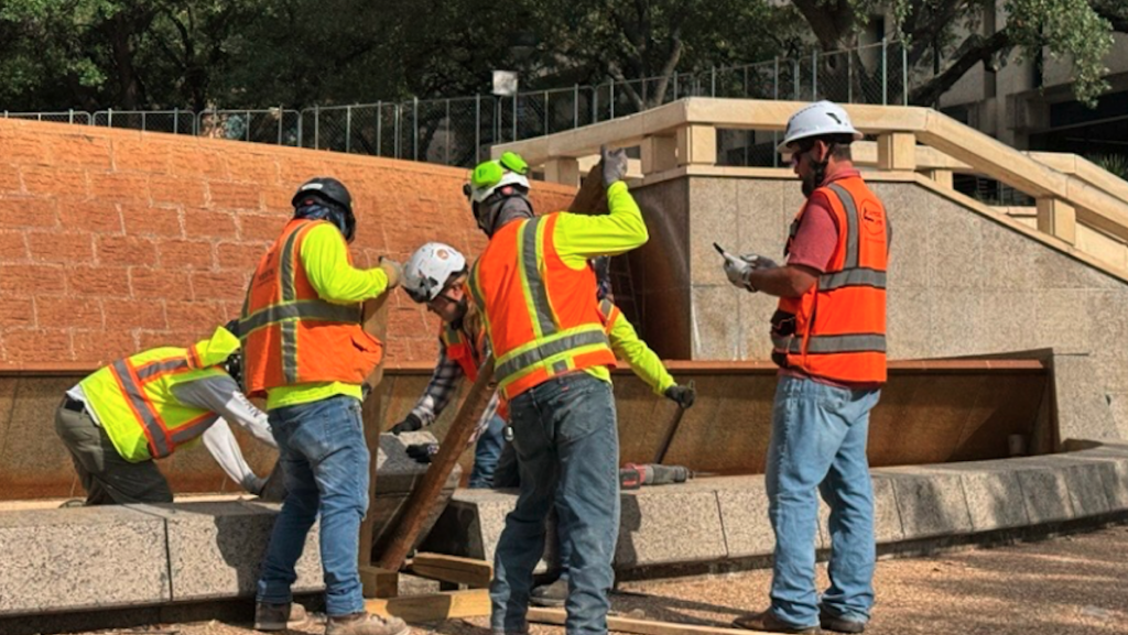Five men in construction protective gear pulling up pieces of granite to demolish an old fountain.